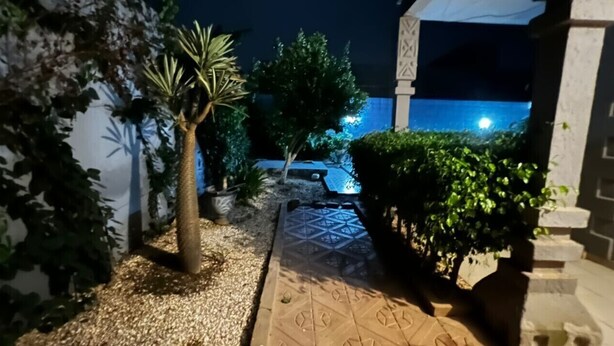 A nighttime outdoor area with a paved walkway, potted plants, and a building in the background. - DIOUF HOUSE (Saly)