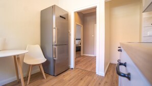 Una cocina compacta con un refrigerador de acero inoxidable, una pequeña mesa blanca con una silla, y piso de madera.