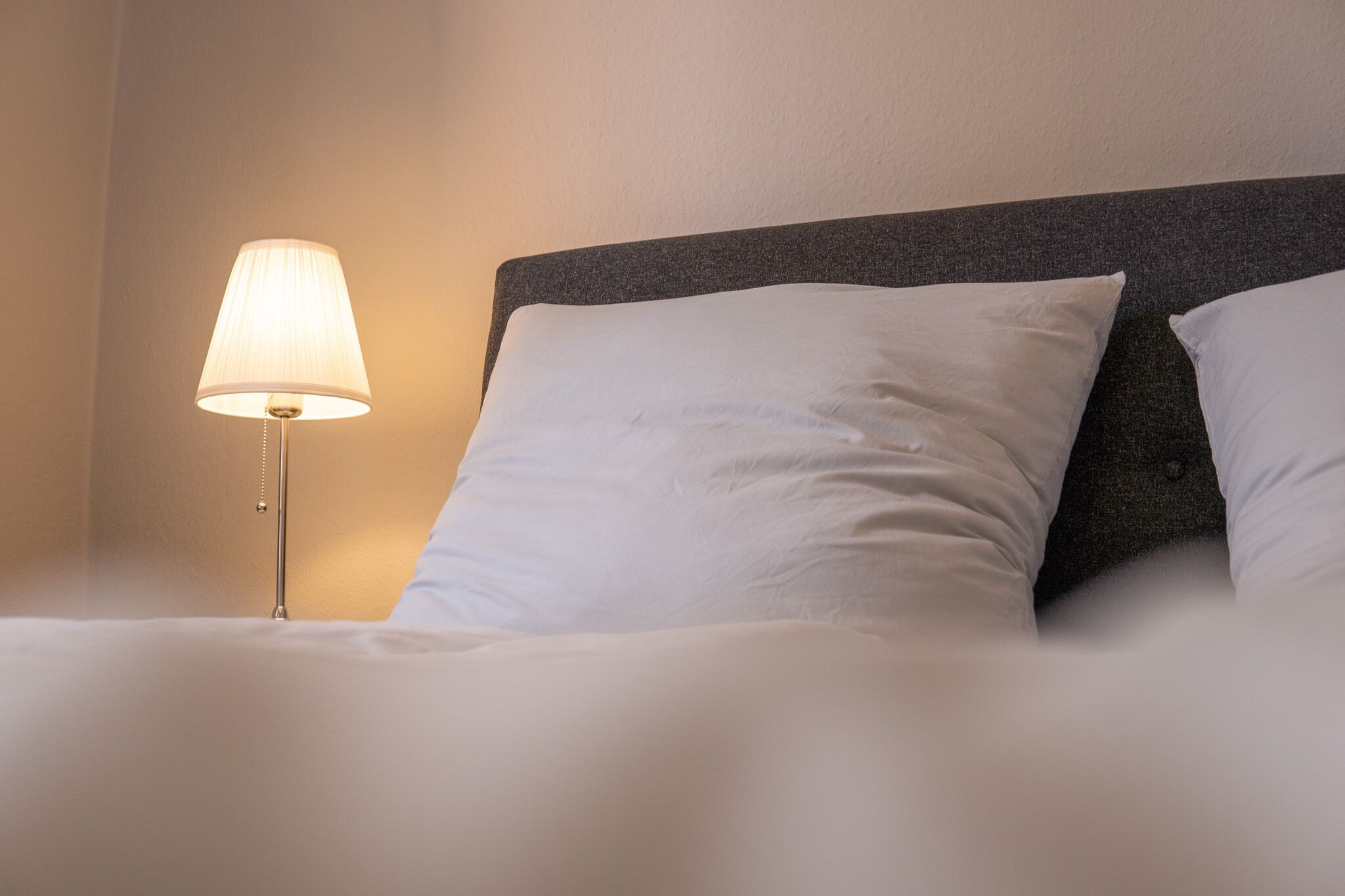 Una cama bien hecha con sábanas y almohadas blancas, una lámpara de noche y un cabecero oscuro.