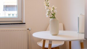 Una mesa redonda blanca con un jarrón blanco lleno de flores blancas, una silla blanca, un radiador blanco y una ventana con vistas a edificios.