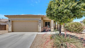 A single-story house with a beige garage door, a small front porch, and a well-maintained garden with various plants and shrubs. - Az morning glory 4bedroom home (Buckeye)