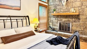Un dormitorio con chimenea de piedra, una cama con cabecera, una mesita de noche con lámpara y una ventana con persianas.