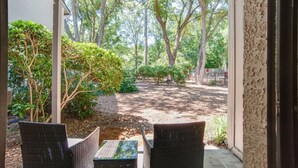 Een overdekt terras met rieten meubilair en uitzicht op een aangelegde tuin met bomen en struiken.