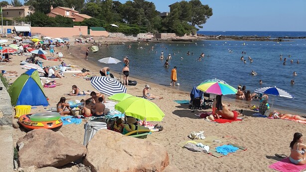 Ein Strand mit badenden, sonnenden und spielenden Menschen. Sonnenschirme, Handtücher und Liegestühle sind vorhanden.