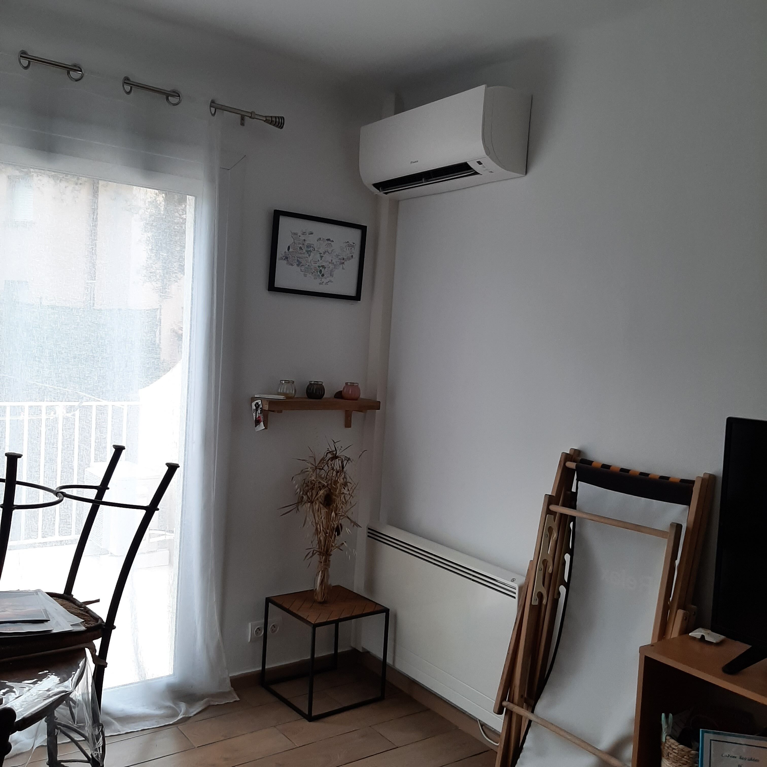 Une pièce avec un climatiseur blanc, un tableau encadré accroché au mur, une petite table en bois sur laquelle se trouve une plante, et une chaise recouverte d’une housse en plastique.