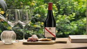Two wine glasses, a bottle of wine, and a small plate with snacks on a wooden table with a blurred green background. - Beachside Studio, TOP NOTCH (MACMASTERS BEACH)