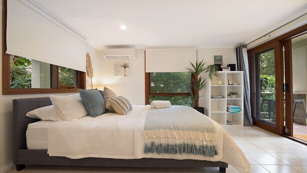 A modern bedroom with a large bed, a bedside table, a bookshelf, and a sliding glass door leading to a balcony. - Beachside Studio, TOP NOTCH (MACMASTERS BEACH)