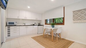 A modern kitchen with white cabinets, a microwave, and a dining area with a table and chairs. - Beachside Studio, TOP NOTCH (MACMASTERS BEACH)