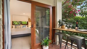 A modern room with a sliding glass door leading to a balcony. The balcony has a wooden table with a bottle of wine, a glass, and a book. There are two bar stools and a potted plant on the floor. - Beachside Studio, TOP NOTCH (MACMASTERS BEACH)