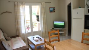A living room with a sofa, a wooden table, and a television. There is a window with curtains, a door, and a refrigerator. - Semi-detached mini-villa in small family residence (San-Gavino-Di-Carbini)