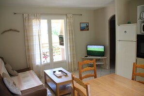Ein Wohnzimmer mit einer Couch, einem Holztisch und einem Fernseher. Es gibt ein Fenster mit Vorhängen, eine Tür und einen Kühlschrank.