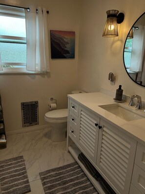 A bathroom with a toilet, sink, and mirror. There is a window with curtains, a radiator, and a painting on the wall. - Winter rental across from the ocean in exclusive San Diego beach community... (Encinitas)
