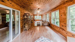 A log cabin interior with a central fireplace, wooden floors, and large windows offering a view of the forest. - Cozy Cabin on the River  (Topton)