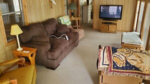 Interior acogedor de una cabaña con un sofá marrón, un televisor, una mesita con una lámpara y una manta estampada en una silla de madera.