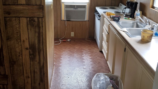 Una cocina compacta con paredes de madera, una ventana con persianas, un fogón, un lavamanos y un cubo de basura.