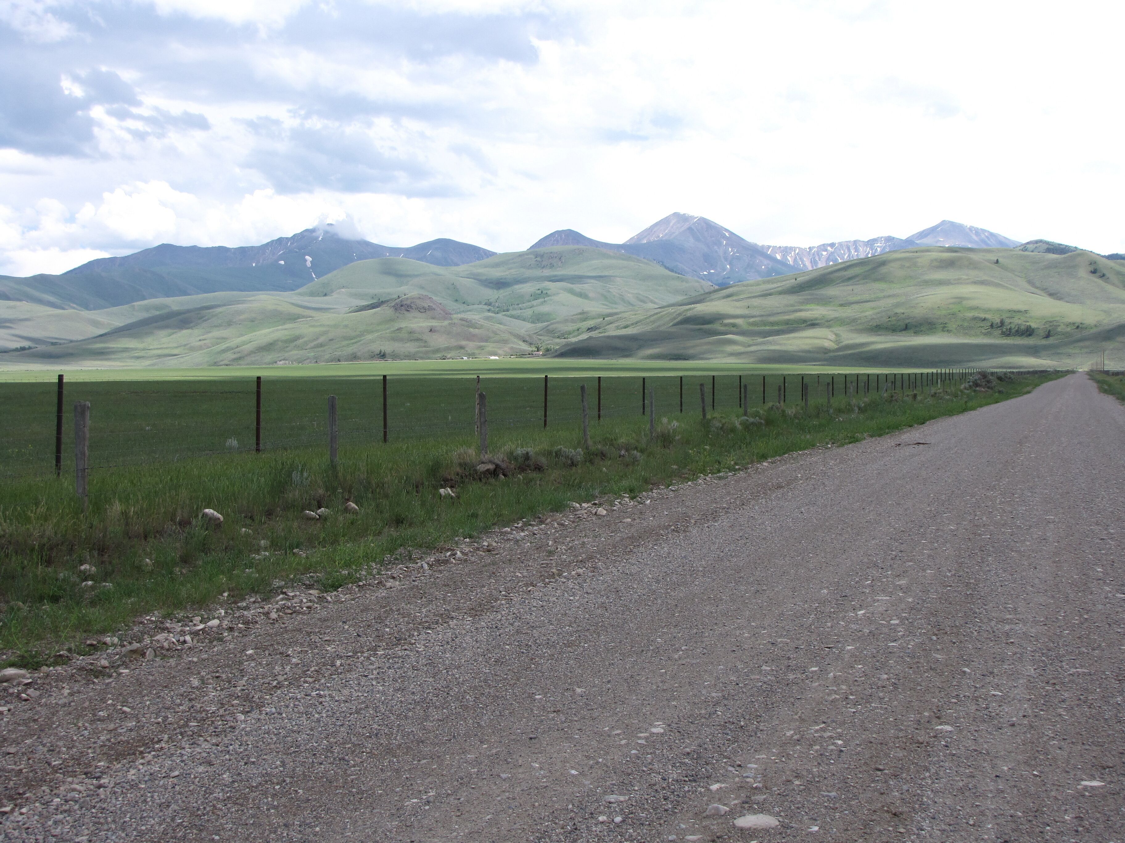 Un chemin de gravier bordé d’une clôture d’un côté et d’un champ d’herbe de l’autre, qui mène vers une chaîne de montagnes.