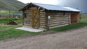 Ein rustikales Blockhaus mit Holztür und Metalldach, gelegen auf einem Schotterweg mit einer Rasenfläche und einem Holzstapel in der Nähe.