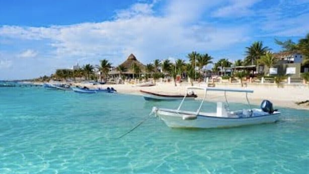 Ein Strand mit klarem türkisblauem Wasser, eine Reihe von Booten und ein Resorthotel mit Palmen und Gebäuden.