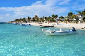 A beach with clear turquoise water, a row of boats, and a resort area with palm trees and buildings.