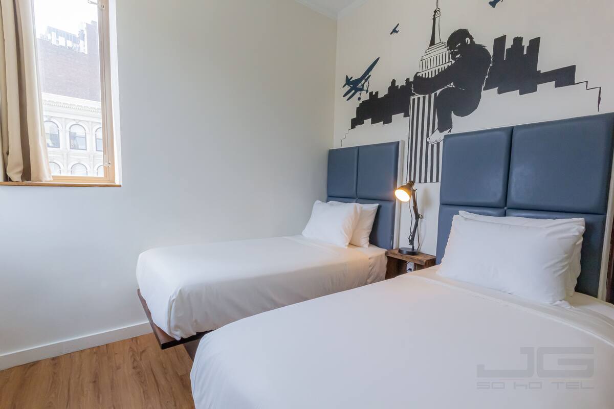 A hotel room with two beds, a cityscape wall mural, and a window with curtains.