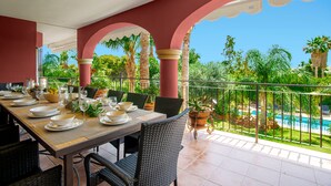 Un comedor con una mesa preparada para cuatro personas, rodeado de sillas de mimbre, con vistas a una piscina y abundante vegetación.