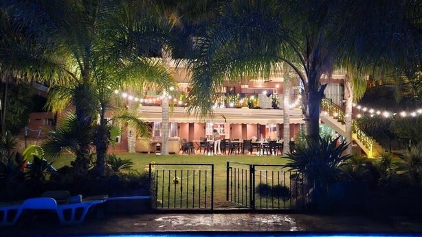 Vista a la piscina de un hotel con entrada protegida, palmeras y luces colgantes.