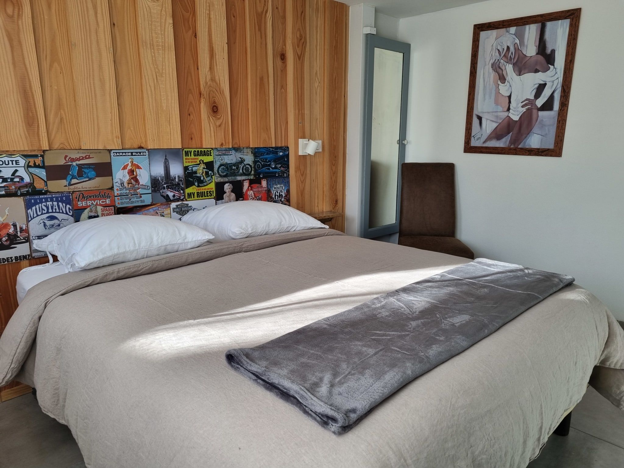 Un lit avec une couverture et des oreillers de couleur beige, une tête de lit en bois ornée d’affiches anciennes sur le thème de l’automobile, un fauteuil marron et un tableau accroché au mur.