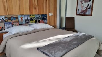 Un lit avec une couverture et des oreillers de couleur beige, une tête de lit en bois ornée d’affiches anciennes sur le thème de l’automobile, un fauteuil marron et un tableau accroché au mur.