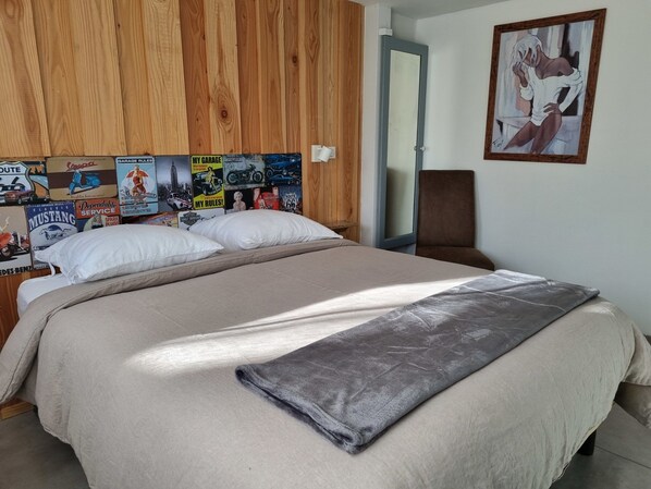 A bed with a beige blanket and pillows, a wooden headboard with vintage automotive posters, a brown chair, and a painting on the wall.