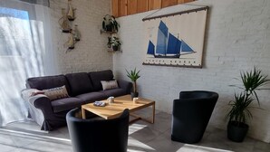 Ein modernes Wohnzimmer mit einer Couch, einem Couchtisch und zwei schwarzen Sesseln. An der Wand hängt ein Gemälde eines Segelboots, und es gibt einige Kübelpflanzen.