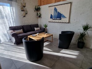 Een moderne woonkamer met een bank, salontafel en twee zwarte stoelen. Op de muur hangt een schilderij van een zeilboot en er staan verschillende potplanten.