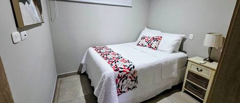 Un dormitorio con cama, una mesita de noche con lámpara, una ventana con persianas y un equipo de aire acondicionado en la pared.