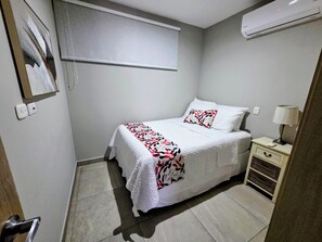 Une chambre à coucher avec un lit, une table de chevet avec une lampe, une fenêtre avec des stores et un climatiseur fixé au mur.