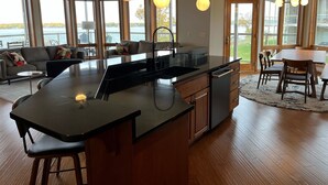 A modern kitchen with a large island, a dining area with a table and chairs, and a living room with a sofa and a view of the outdoors. - Luxurious Lakefront  BB Corner Condo.
Panoramic lake views & 6 waterpark passes. (Arnolds Park)