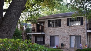 Une maison à deux étages, construite en briques, avec un balcon à l’étage supérieur, une porte de garage et un petit jardin agrémenté d’arbustes et d’un arbre.