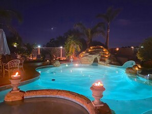 Una piscina nocturna con una formación rocosa y antorchas tiki.