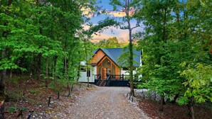 Une cabane au revêtement extérieur en bois, dotée d’un porche et d’un chemin de gravier menant à l’entrée, le tout entouré d’arbres.