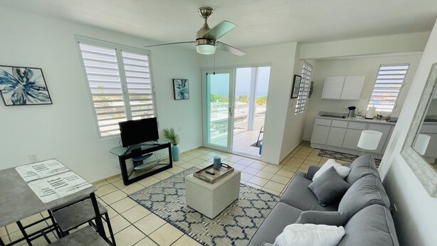 A living room with a grey sofa, a TV on a glass stand, a dining table with chairs, and a kitchen in the background. - Del Mar two separate private suites with ocean views, 5 minutes to beaches (Luquillo)