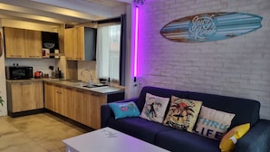 Un espace de vie compact comprenant un coin cuisine, un canapé agrémenté de coussins décoratifs et une œuvre d’art représentant une planche de surf accrochée au mur.