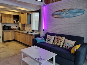 Een gezellig, ruimtelijk ingerichte leefruimte met kitchenette, een bank met decoratieve kussens en een kunstwerk van een surfboard aan de muur.