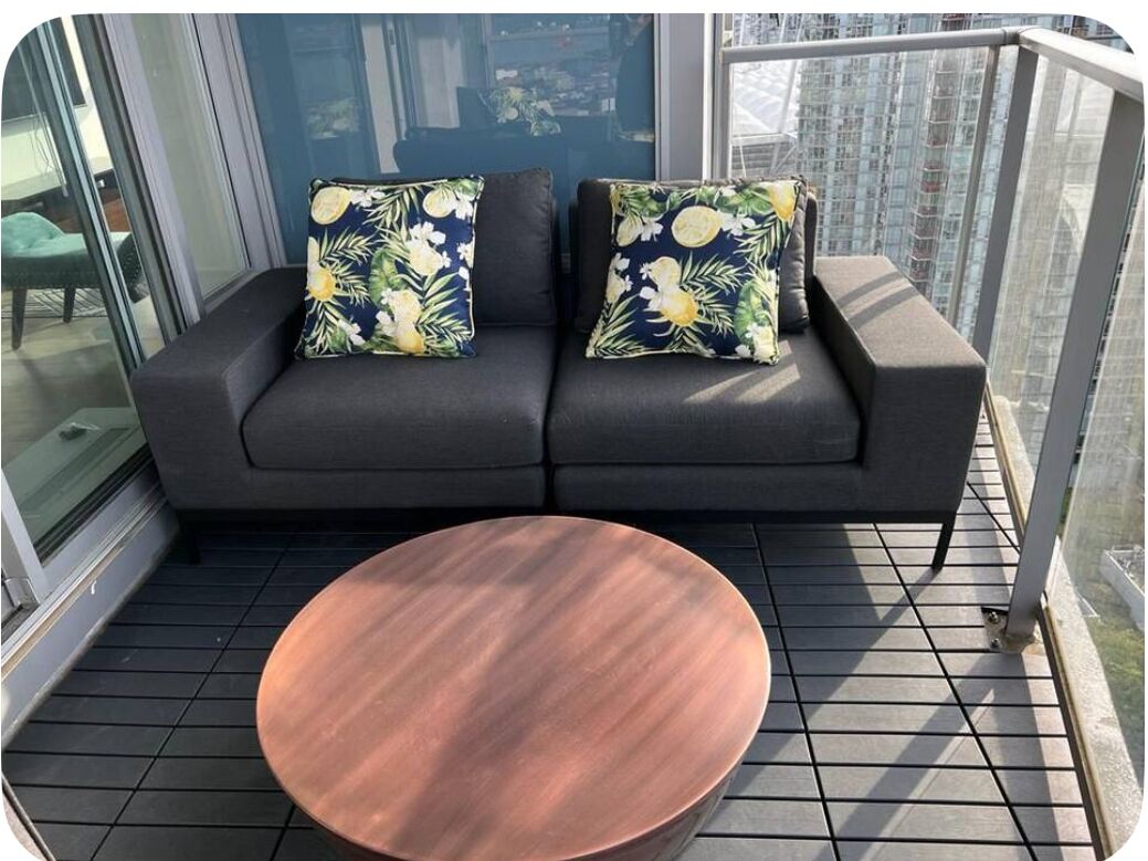 Un balcon avec un canapé noir et deux coussins à motifs floraux, une table ronde en bois et une vue sur la ville.