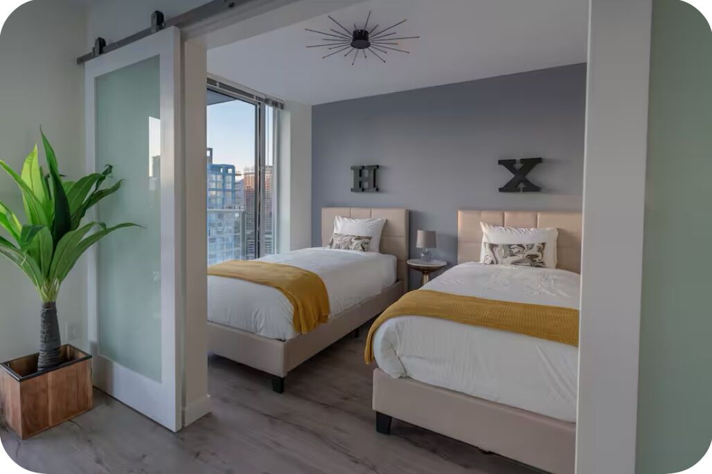Une chambre d’hôtel moderne avec deux lits, une grande fenêtre donnant sur la ville, une plante en pot et un luminaire au plafond au design original.