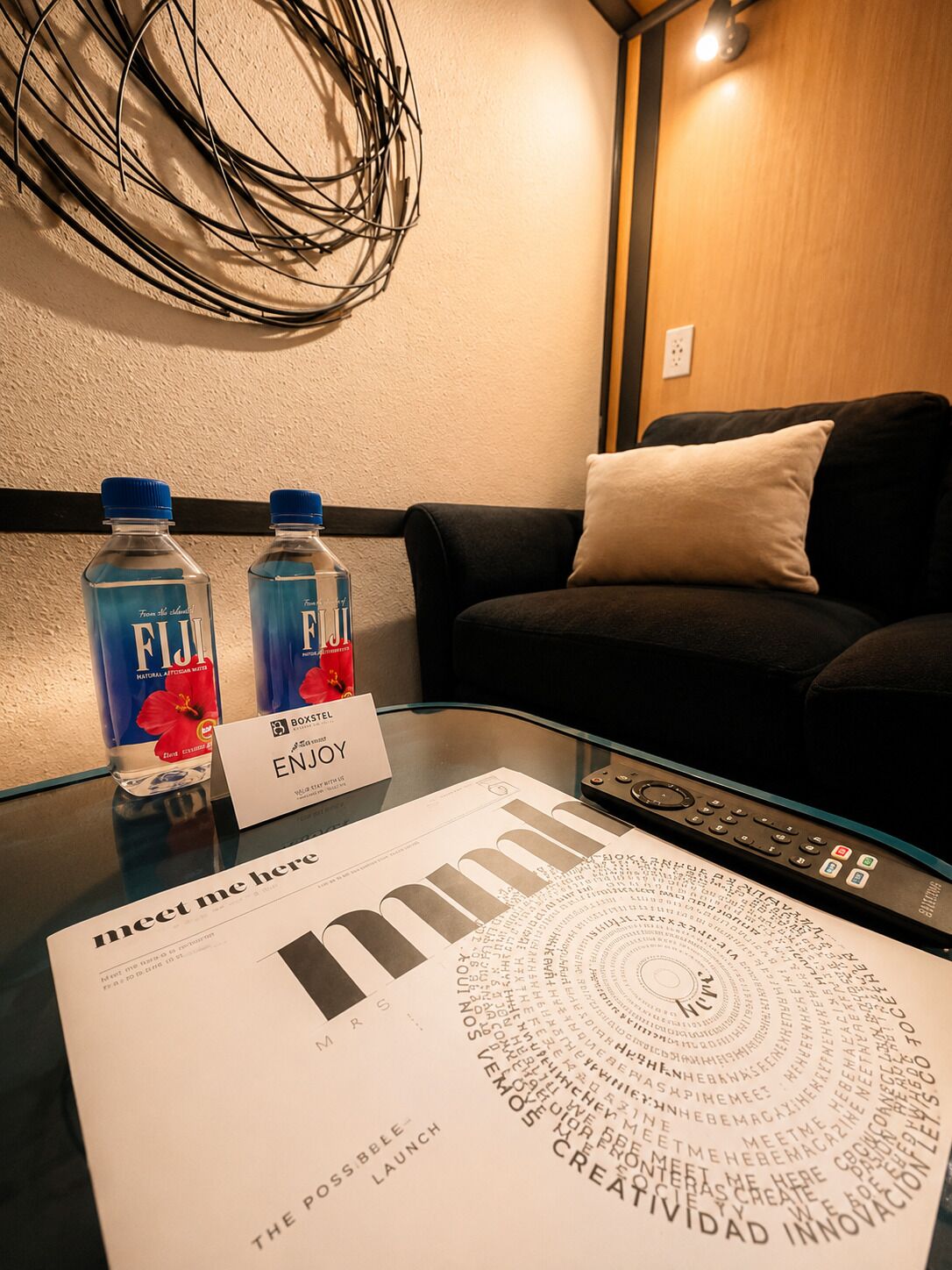 A hotel room with a glass table displaying a magazine, two water bottles, a remote control, and a sign that reads 'meet me here'.