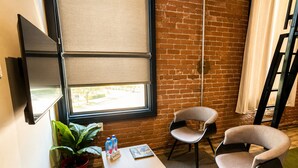 A room with a brick wall, a window with blinds, a flat-screen TV mounted on the wall, a small table with a potted plant and two bottles, and two chairs. - Boxstel - Moderns Stay Hotel (El Paso)