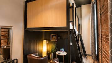 A modern interior with a loft-style bed, leather seating, a small table, and a brick wall.