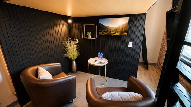 A corner room with two leather armchairs, a small round table with a magazine and water bottles, a potted plant, and a wall-mounted picture. - Boxstel - Moderns Stay Hotel (El Paso)