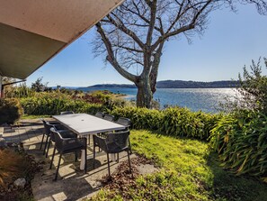 An outdoor patio with a table and chairs, a large tree, and a view of a lake. - Moeroa - Wharewaka Holiday Home (Wharewaka)