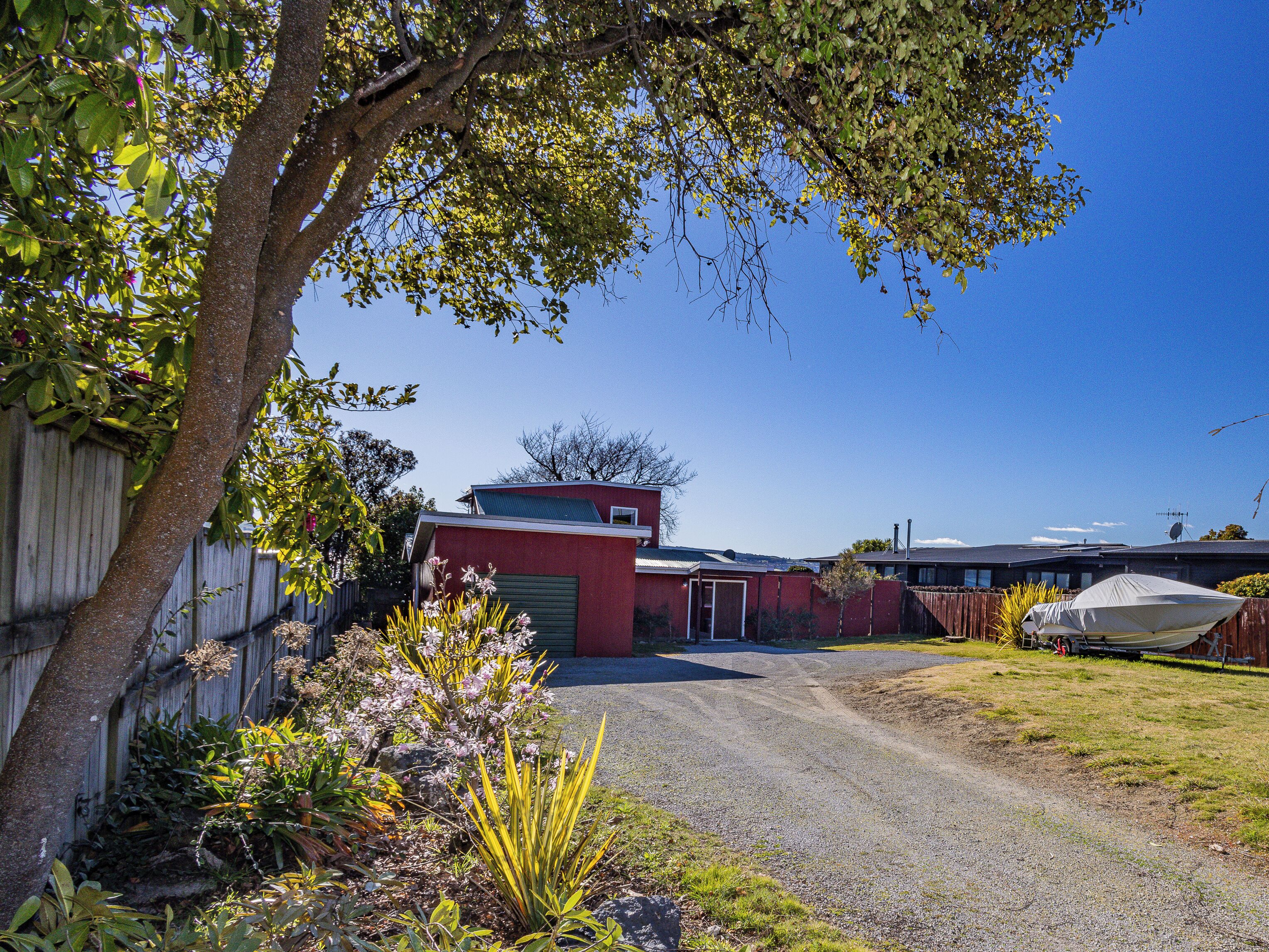 A residential area with a gravel driveway leading to a red garage, surrounded by greenery and a wooden fence.