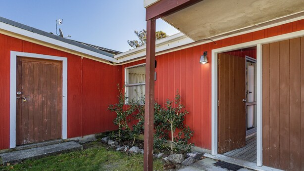 Un edificio rojo con dos puertas de madera, un pequeño jardín con rocas y plantas, y un cielo despejado.