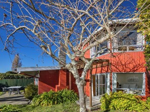 A red building with a glass door entrance, a patio with a table and chairs, and a tree in the foreground. - Moeroa - Wharewaka Holiday Home (Wharewaka)
